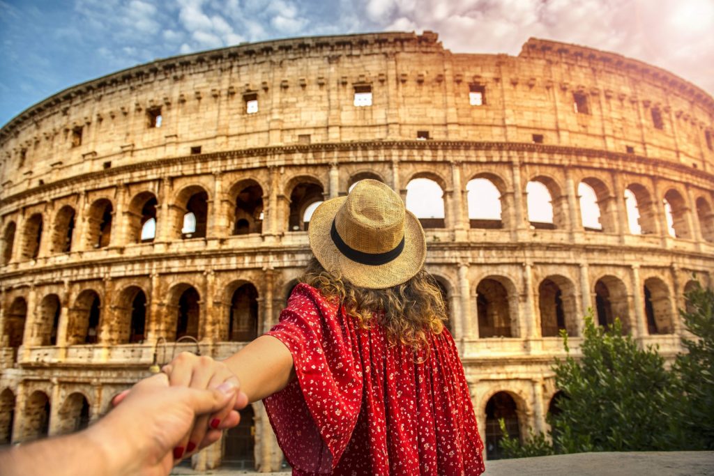 Women in red with a straw hat faces the Colosseum in Rome while hoding the hand of a person behind the camera
