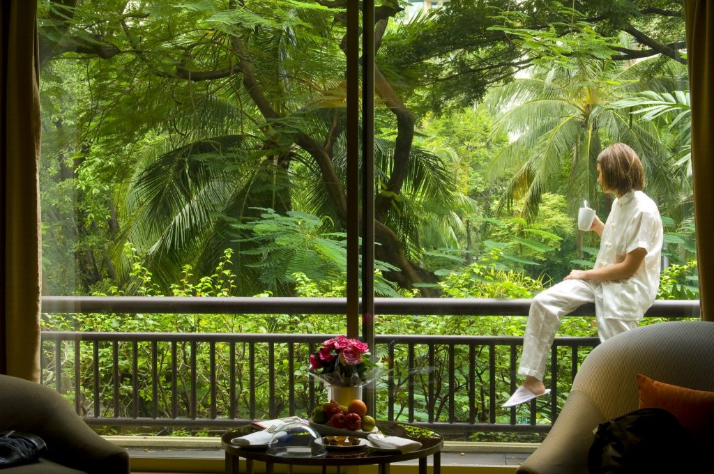 a woman sits on the railing of an eco-friendly accomodation starting at the plam trees and greenery
