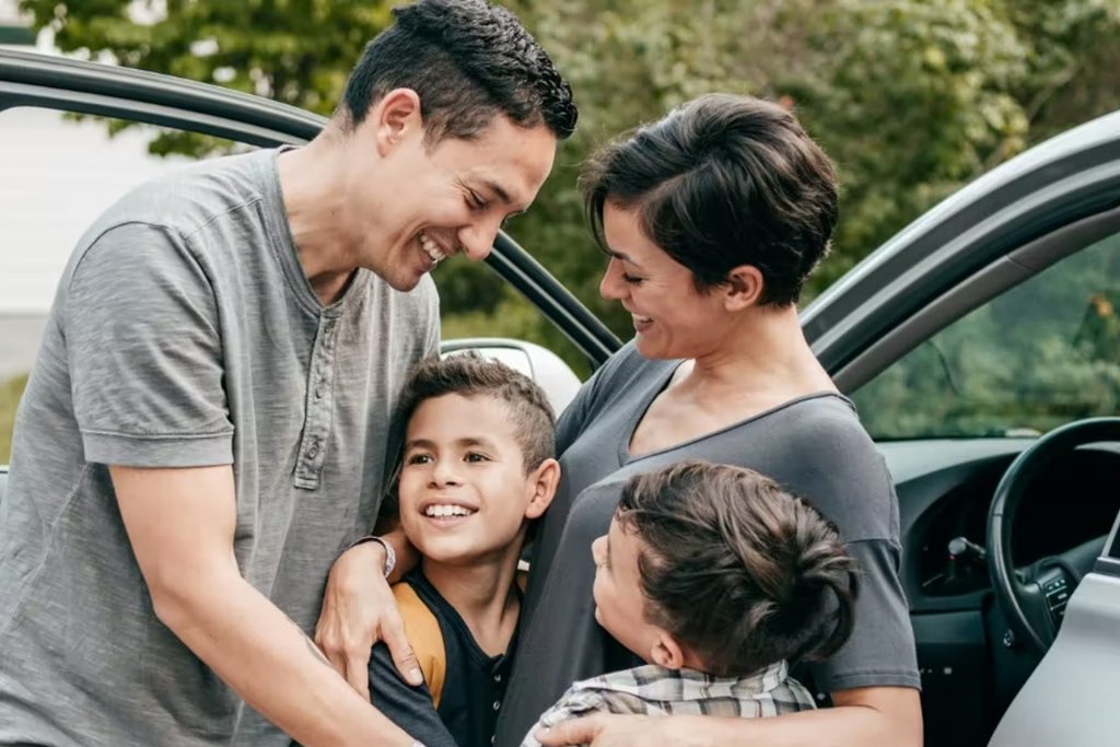 A young family with two kids hug beside an open car door