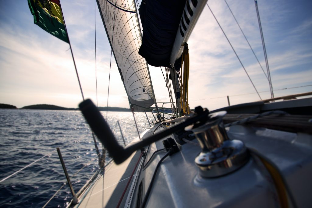 The deck of a sail boat, sailing calm waters towards shore on the horizon