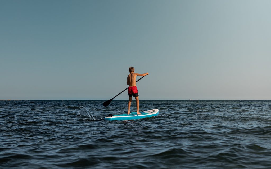 A boy in red shorts makes a spalsh with the paddle as he paddles towards the summer horizon on a blue stand up paddle board