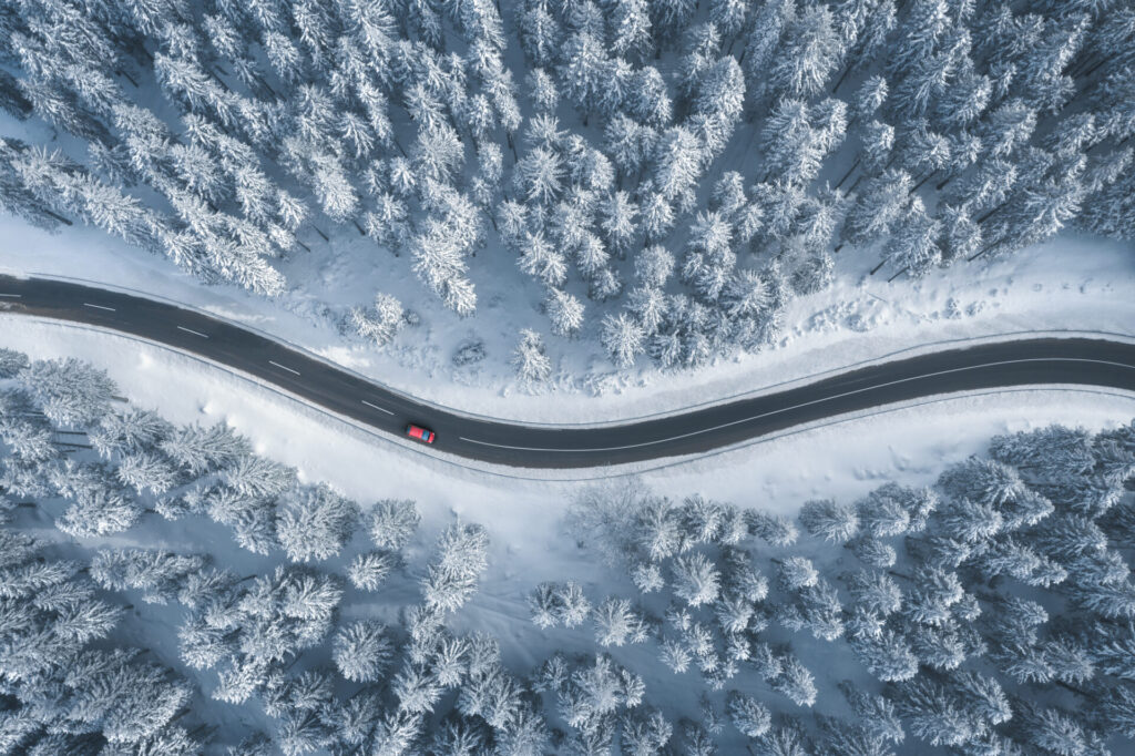 Red car driving on black asphalt road leading through snowcapped winter forest.