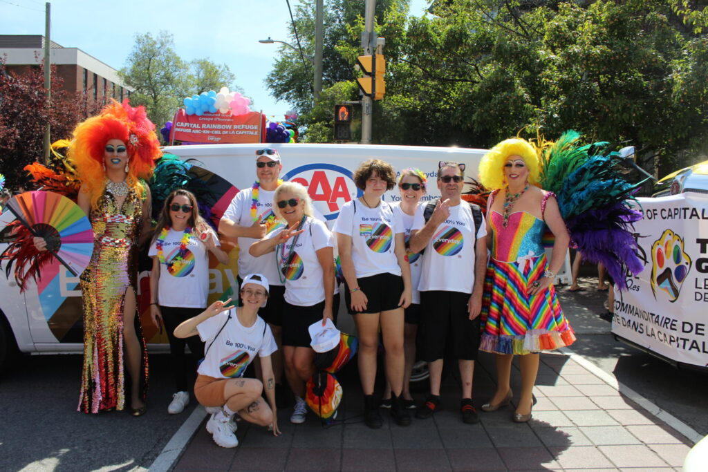 A group in white tsirts pose with drag queens infront of the CAA Everyday Van at Ottawa pride parade