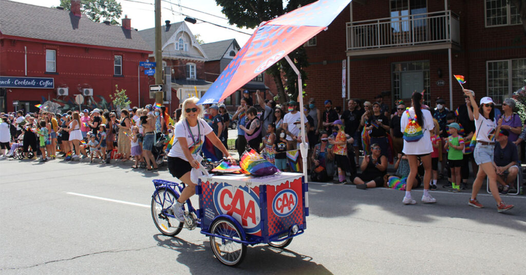 Woman on a bicycle with freezer and umbrella peddals through Ottawa Pride celebrations