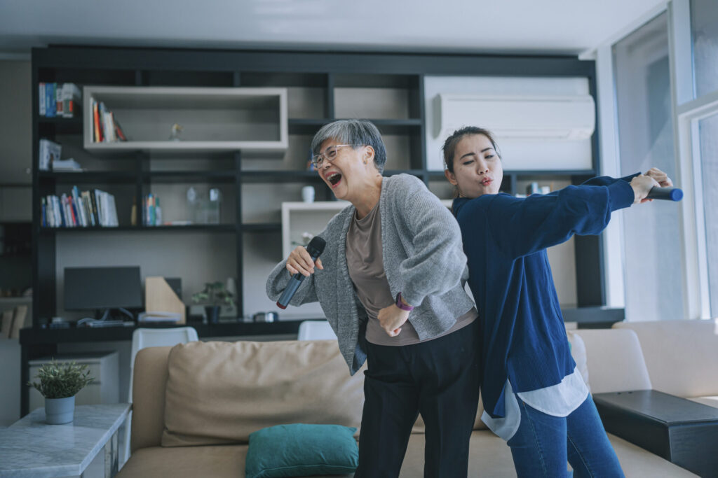 A senior woman singing karaoke and dancing with her daughter in the living room during weekend leisure activities