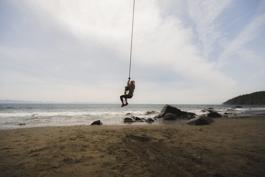 A young blonde woman swings on a rope swing at the scenic Mystic Beach located along the shores of the Strait of Juan de Fuca on Vancouver Island, British Columbia, pacific northwest Canada.