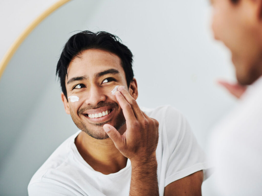 Man applying face skincare cream while grooming looking at the mirror in the bathroom in his morning routine at home. Close up portrait of handsome guy using facial lotion for clean and clear skin